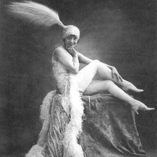 Black and white photograph of Moulin Rouge Burlesque star, Mistinguett, posing on a pedestal in a feather-edged gown and hat.