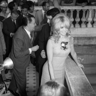 Seen from above, Brigitte Bardot, in a short dress with shaggy blond hair, smiles during a busy outside event at the hotel.
