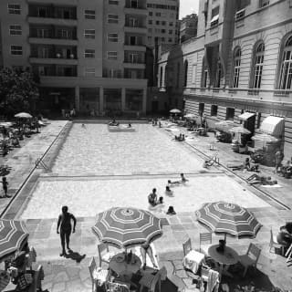 Timeless monochrome shot of the pool, with three parasols and tables, and people lunching and lounging at the poolside.