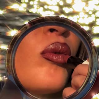 The lower face of a woman applying red lipstick is caught in a round compact mirror, with sea behind, taken by Sam Youkilis.