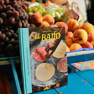 The El Bajío cookbook, with a cover photo of four heated tortillas, rests on a blue market table by an abundant fruit stall.