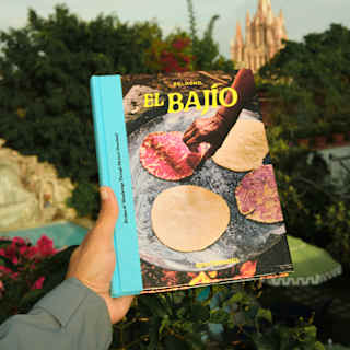 A hand holds the El Bajío book, with plain and red tortillas on the cover, in front of a view of San Miguel's iconic church.