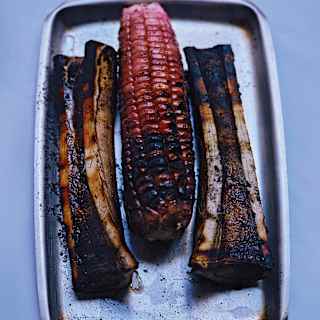A metal tray holding a grilled cob of red corn placed between two pieces of charred, grilled bone marrow, all arranged neatly on a white background.