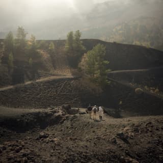 In a low-light image, three walkers hike the undulating, black rocky paths in cloudy Parco dell'Etna, seen from behind.