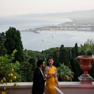 A woman in a yellow dress talks with a man in black jacket on Grand Hotel Timeo's terrace overlooking the Taormina coast.