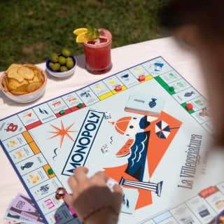 At a table with a red cocktail, olives and crisps, a guest plays Monopoly, the La Villeggiatura edition, seen from above.