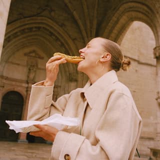 A woman in a beige coat eats a nut-covered chocolate éclair straight from a patisserie paper by the steps of a church.