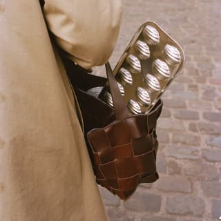 A silver-tone baking tray for making Madeleines juts from the leather bag of a woman in a beige coat, seen from behind.