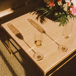 Two Champagne flutes create long shadows across a low table with a flower arrangement, seen from above in evening light.