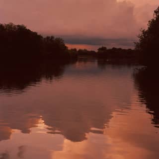 Burgundy clouds bloom in a purpling sky, reflecting the sunset spectacle on the serene waters of tree-lined river below.