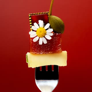 Artistic food image of an upright fork, prongs spearing a cube of cheese, topped with a fruit jelly, daisy flower, caper and gem.