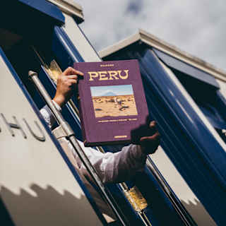 Low-angle image looking up at a train carriage doorway where a man's arms hold out a copy of the Belmond publication, Peru.