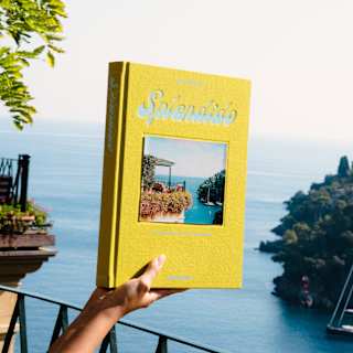 A hand holds a copy of Splendido, with a yellow-bordered cover-photo of a Portofino bay view, replicated in this very image.