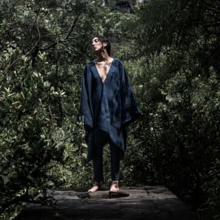 A barefoot female model poses in a black poncho with a deep v-neck by Collectiva Concepció, worn over black trousers.