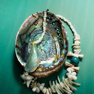 A white shell-fragment bracelet with nacra mussel rests on a mother-of-pearl abalone shell, placed on a green surface.