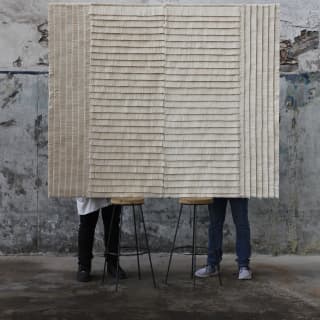 The legs of two men are visible behind a raised Estrella Wall Rug designed by Caralarga using repurposed raw cotton thread.