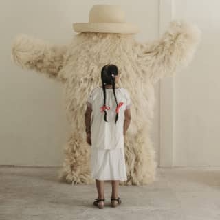 Seen from behind, a girl with long braids looks at a white fluffy monster designed by Fernando Laposse using agave fibres.