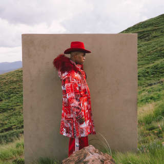 In a hillside magazine fashion shoot, a male model wears a red print jacket with feather trim and Stetson by Thebe Magugu.