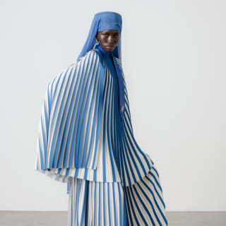 A model wears a blue and white striped plissé skirt and cape-like top by Thebe Magugu, with white heels and a blue headdress.
