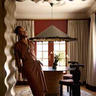 A person in a brown dress leans against a wavy-edged archway in a warmly lit room with a round table, black chairs, a unique lampshade, and long beige curtains covering the windows.