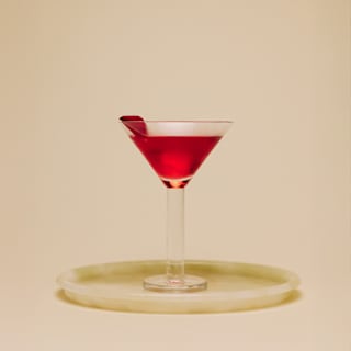 Eye-level view of a glowing, deep red Rose Martini cocktail, garnished with a petal, served in a thick-stemmed Martini glass.