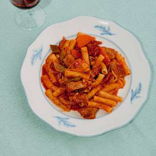 Looking down on a plate of rigatoni pasta in a rich vegetable ragout, served with a glass of red wine on a pale blue tablecloth.