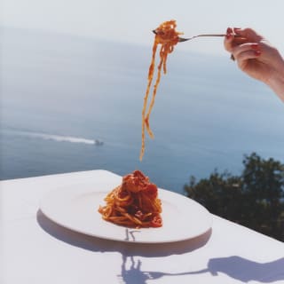 Image from the Liguria cookbook shows a bowl of three-tomato Spaghetti alla Elizabeth Taylor enjoyed at an ocean-view table.