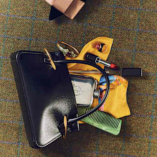 A black handbag is open on a checked surface, revealing a yellow glove, green comb, perfume bottle, sunglasses, and a charging cable. A person wearing grey shoes with a pink bow is partially visible at the top.