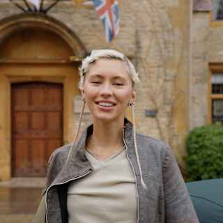 Actress Iris Law, with blond hair and two long thin plaits, smiles to camera outside Le Manoir's front door, in soft-focus.