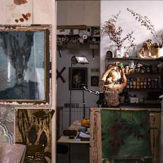 A cosy, cluttered artists' studio with paintings, art supplies, plants, and dried flowers. Works of art and reference photos cover the walls. A desk lamp and shelves of jars are visible in the background.