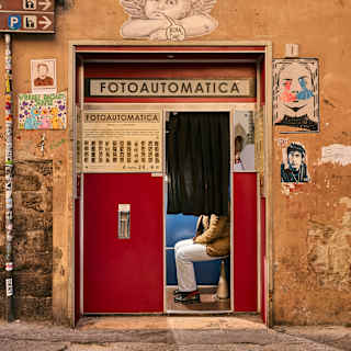 A person sits inside a red photo booth with a black curtain, set in an old, orange wall decorated with graffiti, posters, and a white angel illustration above the booth labelled Fotoautomatica.