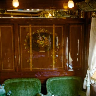 Two plush green chairs sit beneath a floral marquetry panel, seen over a table filled with glasses in the Etoile du Nord car.