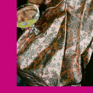 A woman holds a drink on the lap of her rose silken skirt with floral metallic brocade, pictured close-up on pink background.
