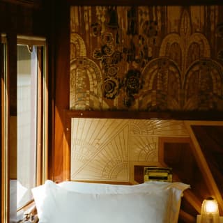A single bed in crisp white linen with a propped pillow settles between a cabin window and a side table with a glowing lamp.