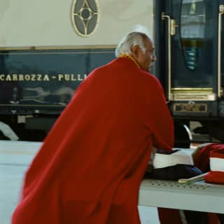 A grey-haired man sits on a platform bench in front of the train, in a long red cloak with gold piping, seen from behind.