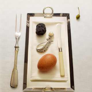 Looking down on a tray with an antique knife, egg, black truffe and vintage brooch at a table with a pickle fork and caper.