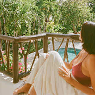 A woman in a red top and white skirt sits on her balcony above a garden with lush palms, walkways and a swimming pool.
