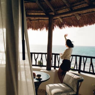 A floating drape partly obscures a sea-view balcony where a woman stands at the railings behind a cushioned chair and waves.