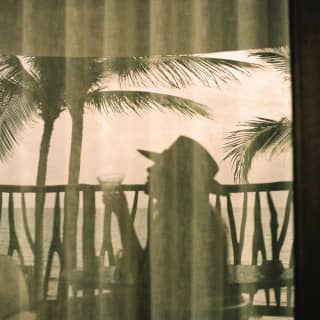 A man in hat enjoys a drink on his suite balcony, silhouetted against the peach sky and viewed through diaphanous drapes.