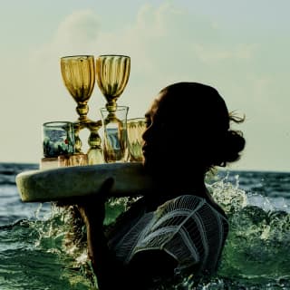 A woman carries a round tray of drinks and unusual glasses on her right shoulder as she wades through waist-high ocean water.