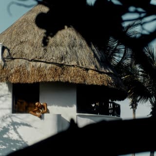 A woman leans on the white-painted stairwell at the upper entrance of her thatch-roofed suite, seen through tree branches.
