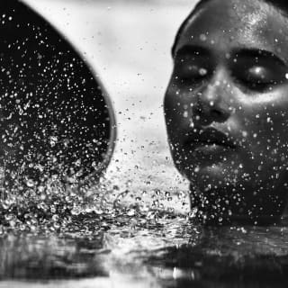 A close-up of a woman in water up to her neck, holding a bowl, obscured by droplets, captured in monochrome by Jack Davison.