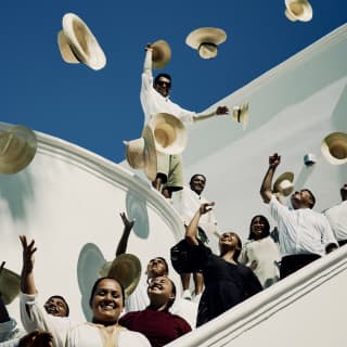 Smiling male and female members of Maroma's staff crowd onto a white stairwell, tossing Panama hats into a deep blue sky.
