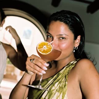 A dark-haired woman in an off-the-shoulder green dress sips from an orange-garnished cocktail at  Bambuco.