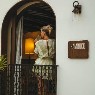 Bambuco's wood sign adorns the exterior wall by an open arched window where a blond woman in a white dress sips from a glass.
