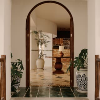 Potted monstera plants guard the archway entrance to Woodend with a wooden door frame and glazed green-tile threshold.