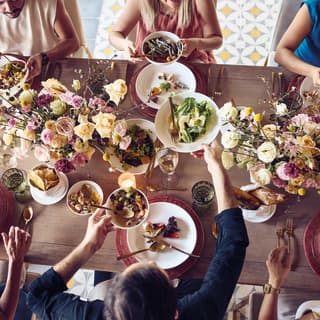 View from above of a special occasion meal as six guests pass zesty-looking bowls of food between garden-flower centrepieces.