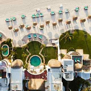 Bird's eye view of beachfront villas with turquoise pools and lawns adjacent to a white beach dotted with chairs and parasols.