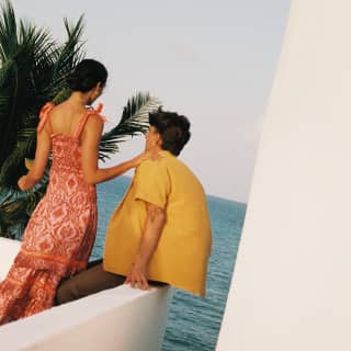 A woman in red dress stands by a man in a yellow shirt, sitting on the edge of the roof terrace overlooking the rolling sea.