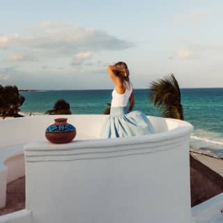 A woman in a white top and blue skirt stands at Maroma's El Mirador circular white-walled viewpoint, gazing at the azure sea.